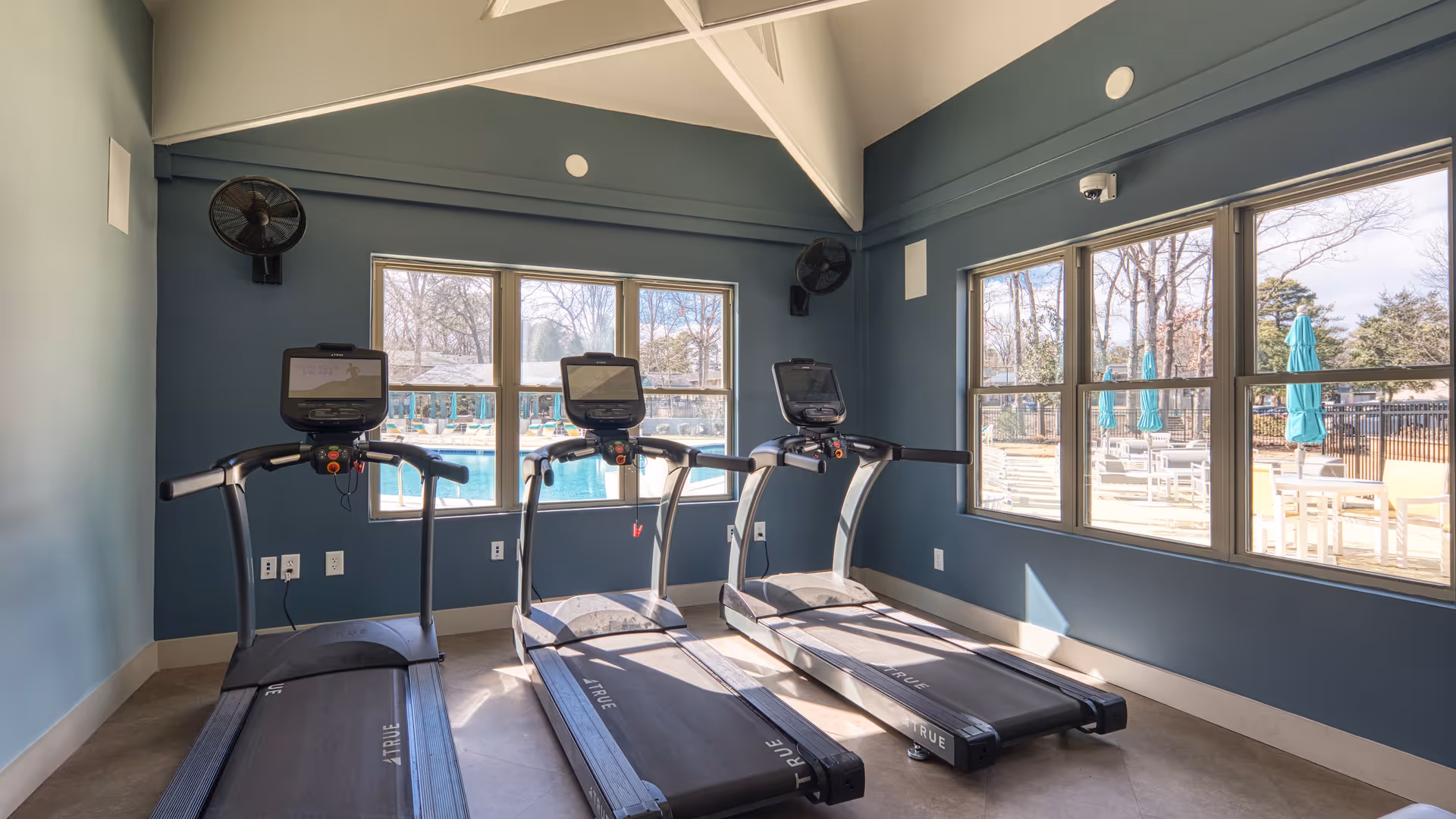 A small fitness room with three treadmills facing large windows that overlook an outdoor pool area with lounge chairs and umbrellas. The walls are painted blue, and the ceiling has white beams.
