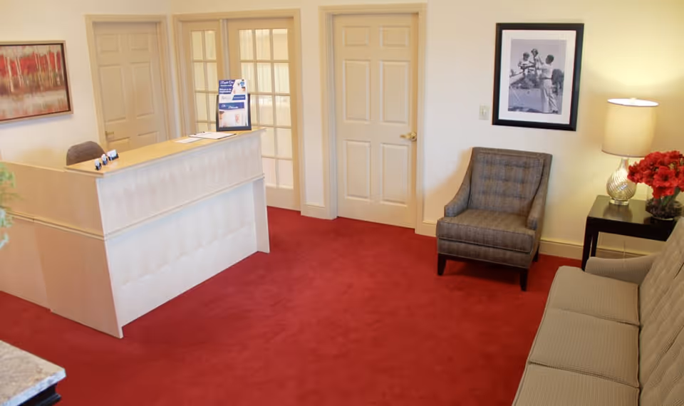 Reception area with a light wood reception desk, a gray upholstered chair, a gray sofa, a side table with a lamp and red flowers, a framed black and white photo on the wall, and a red carpeted floor.