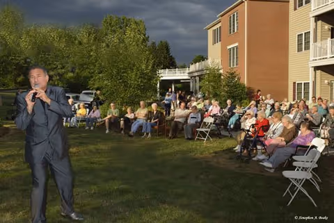 A male performer sings for a seated outdoor audience of seniors on the lawn beside a multi-story residential building.