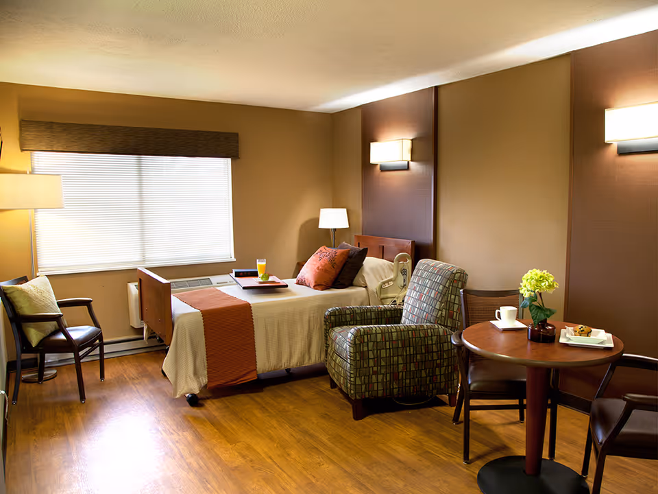 A cozy bedroom in a healthcare facility with a single bed made up with beige bedding and an orange throw. There is a tray with a glass of orange juice on the bed. Next to the bed is a patterned armchair and a small round wooden table with a flower vase, a cup, and a plate with a muffin. The room has wooden flooring, beige walls with brown accent panels, a window with blinds, and several light fixtures including a floor lamp and wall sconces.
