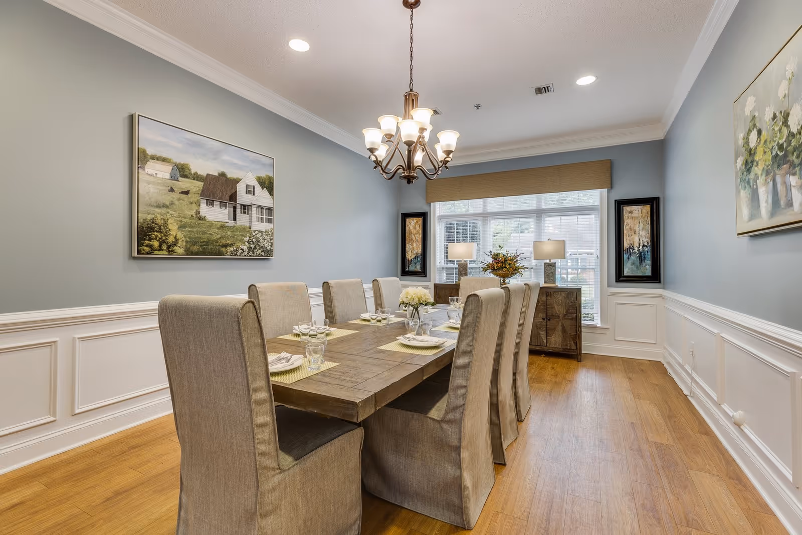 A well-lit dining room with a wooden dining table set for eight people. The chairs are upholstered in beige fabric. The room has light blue walls with white wainscoting and wooden flooring. A chandelier hangs above the table. There are two framed paintings on the walls and a sideboard with two lamps and a floral arrangement in front of a large window with blinds.