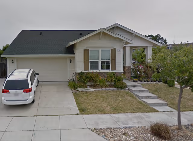 Single-story house with a gray roof, beige siding, and brown shutters. There is a white minivan parked in the driveway. The front yard has a small lawn, some bushes, and a tree near the sidewalk. Concrete steps lead up to a covered front porch.