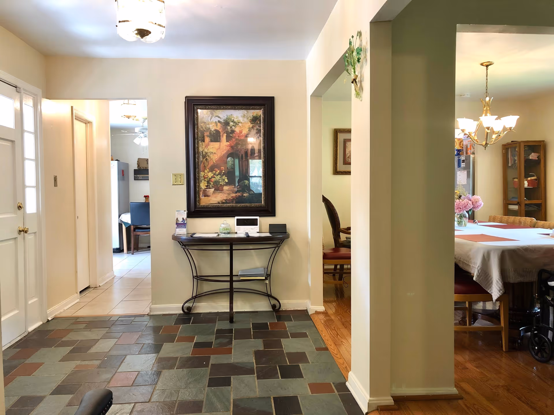 Interior view of a senior living facility showing a tiled entryway with a small table against the wall holding a few items and a framed painting above it. To the right, there is a dining area with a table covered with a white tablecloth, chairs, a chandelier, and a vase with pink flowers. The kitchen is visible in the background through an open doorway.