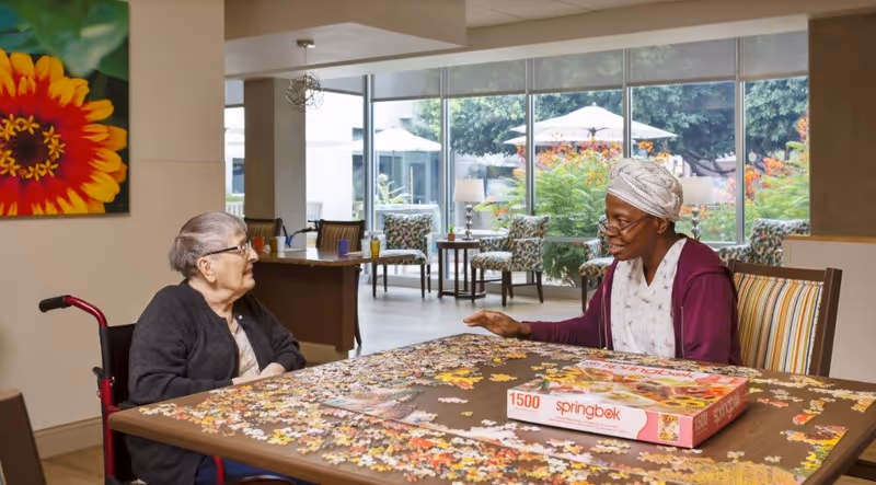 Two elderly women sitting at a table working on a 1500-piece Springbok jigsaw puzzle in a bright room with large windows showing outdoor greenery and patio umbrellas. One woman is in a wheelchair and the other is wearing a headscarf and glasses.