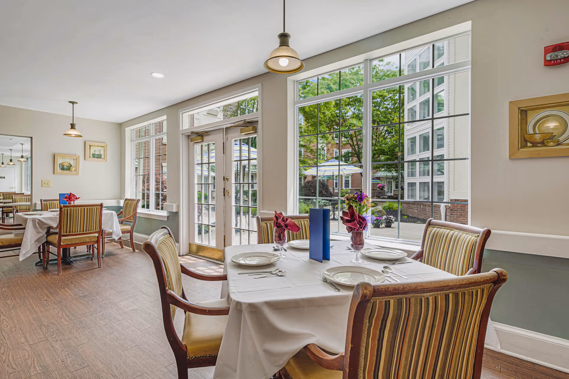 A bright dining room with large windows and glass doors that open to an outdoor patio. The room features tables covered with white tablecloths, set with plates, utensils, and glasses with folded red napkins. Striped upholstered chairs surround the tables, and pendant lights hang from the ceiling. Outside, umbrellas and greenery are visible.