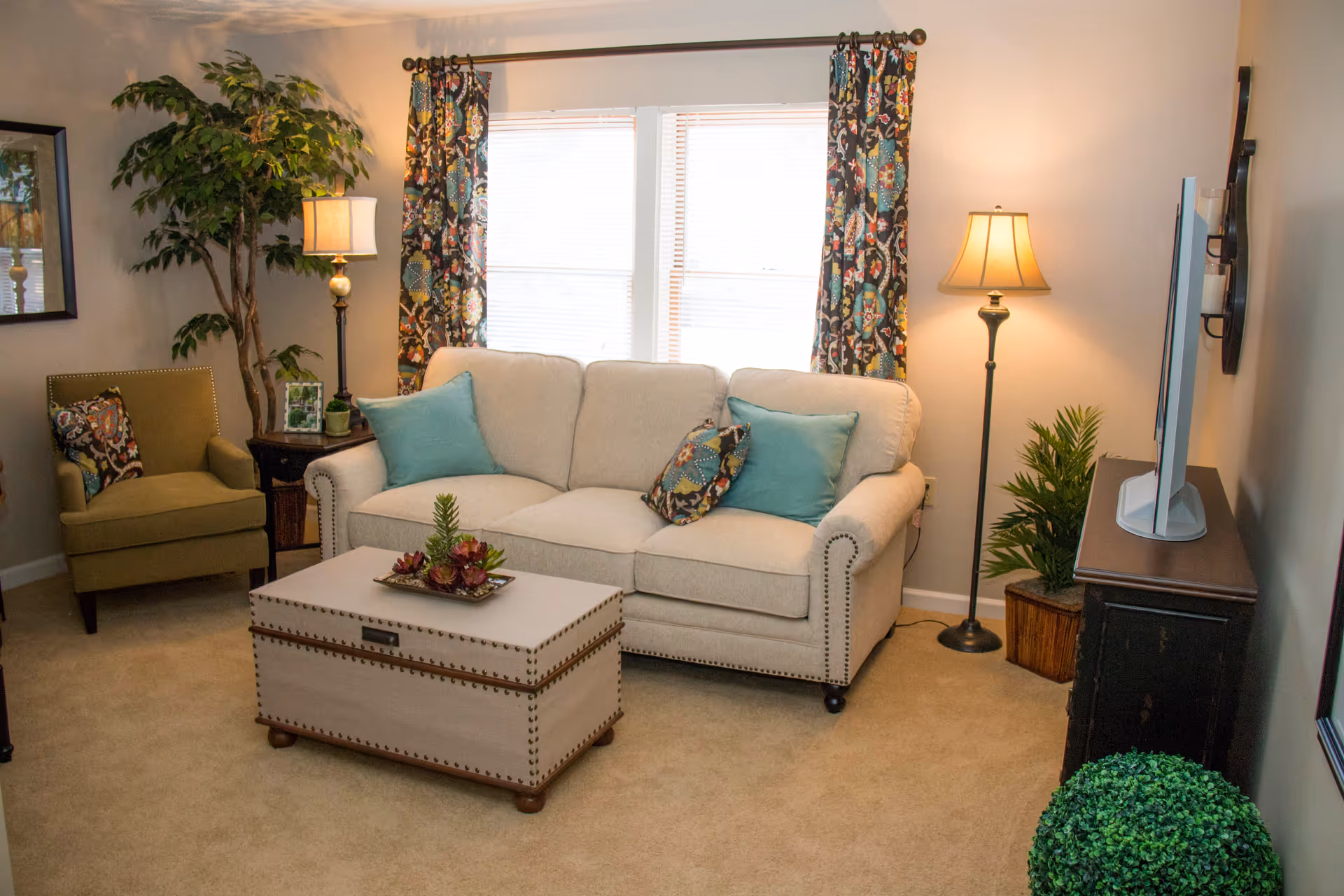 A cozy living room with a beige sofa adorned with blue and patterned throw pillows, a matching armchair with a patterned pillow, a wooden coffee table with a decorative plant centerpiece, two floor lamps, a TV on a dark wooden stand, and several green plants. The room has beige carpeting and a window with colorful patterned curtains.