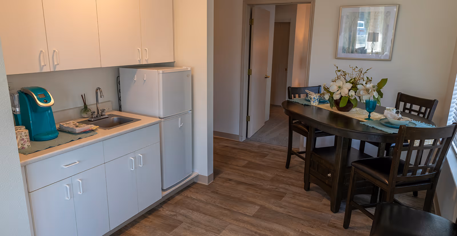 A small kitchen area with white cabinets, a countertop with a sink, a teal coffee maker, and a white refrigerator. Adjacent to the kitchen is a dining area with a dark wooden table set with floral decorations, cups, and plates. There are four dark wooden chairs around the table, and a framed picture hangs on the wall above. The floor is wood-patterned, and a hallway with open doors is visible in the background.