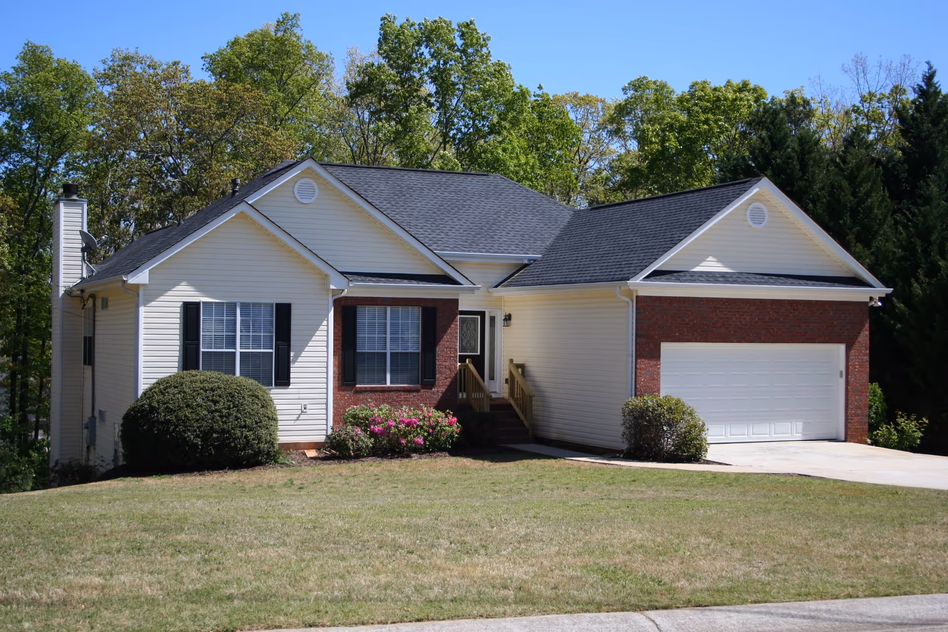 Front exterior view of a single-story house with white siding and red brick accents, a black shingled roof, a two-car garage, and a small front porch with steps. The house is surrounded by green bushes and trees under a clear blue sky.