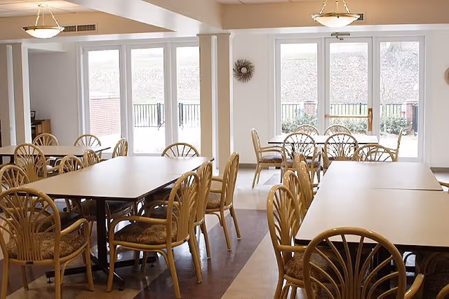 A bright dining room with multiple rectangular tables and wooden chairs with cushioned seats. Large windows and glass doors let in natural light and offer a view of an outdoor area with a railing and greenery. The room has light-colored walls and ceiling lights.