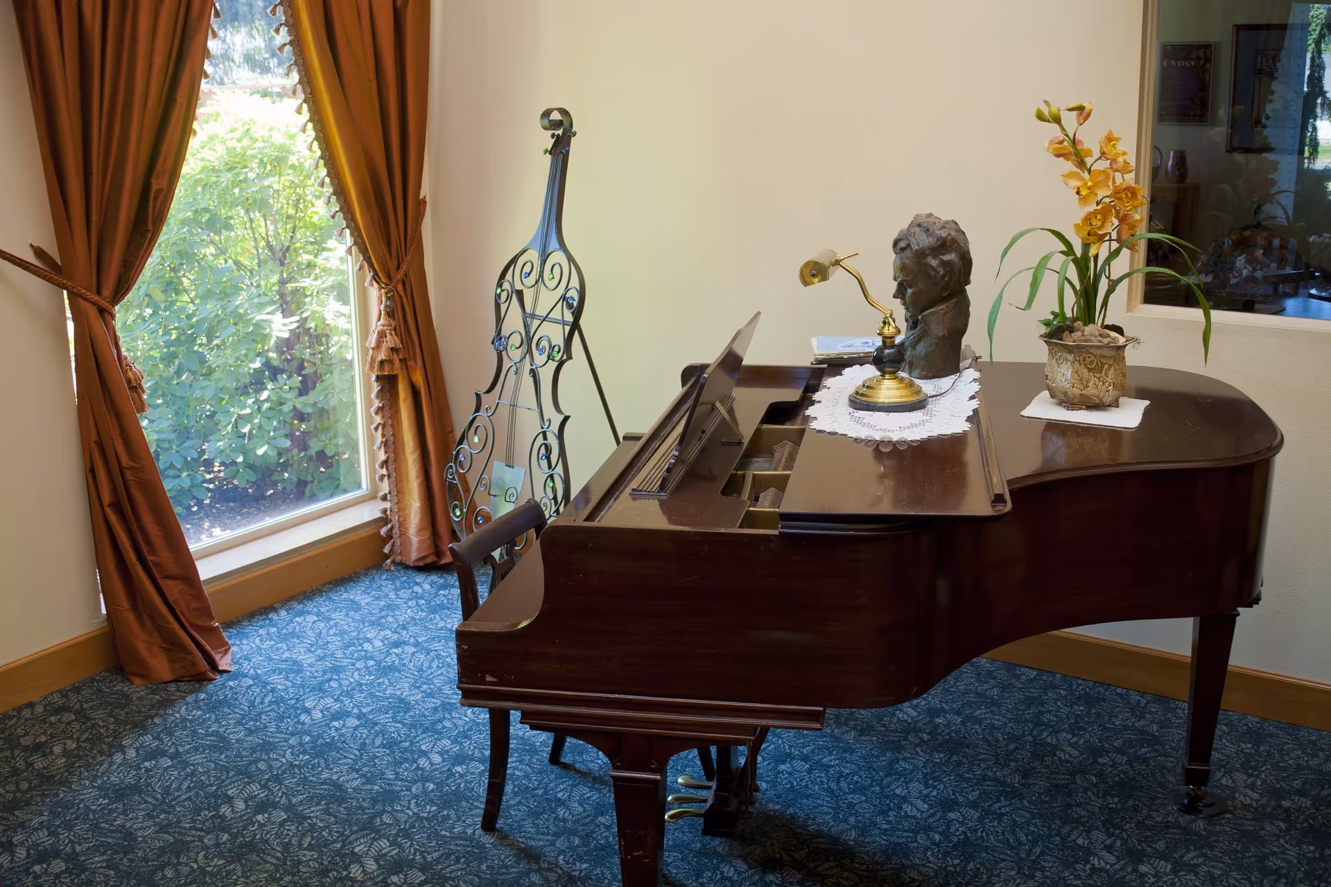 A grand piano in a carpeted room by a window with brown curtains, topped with a small lamp, a bust sculpture and a potted plant.
