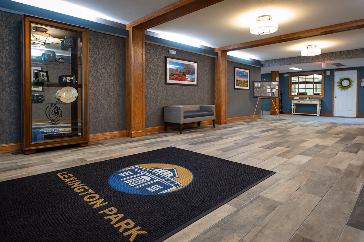 Spacious reception lobby with a branded 'Lexington Park' entrance mat, seating bench, display cabinet, framed artwork, and a distant reception table.
