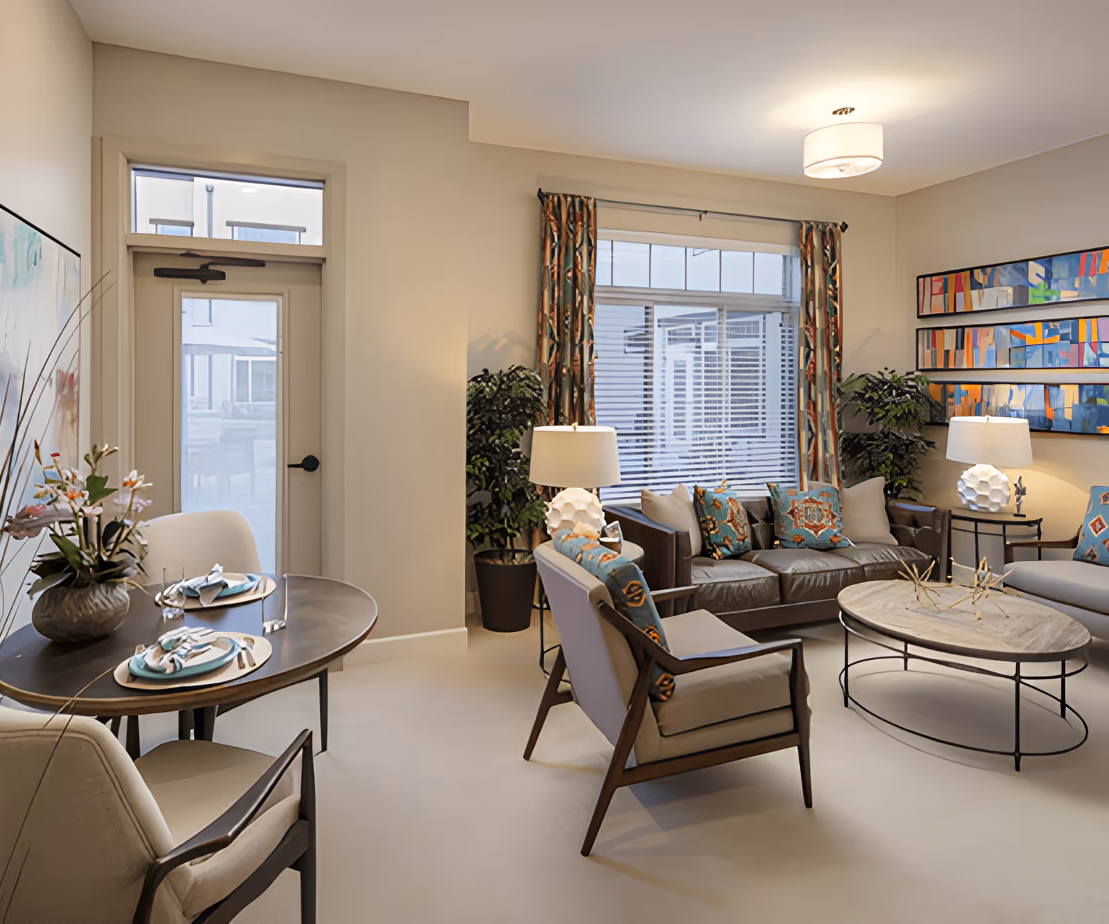 A cozy living room and dining area in an assisted living facility. The living room features a brown leather sofa with colorful patterned pillows, two armchairs, a round coffee table with decorative items, and two table lamps with white shades. Large windows with patterned curtains allow natural light to fill the room. Adjacent to the living room is a small dining table set for two with plates, napkins, and silverware. The walls are decorated with abstract artwork, and there are potted plants adding greenery to the space.