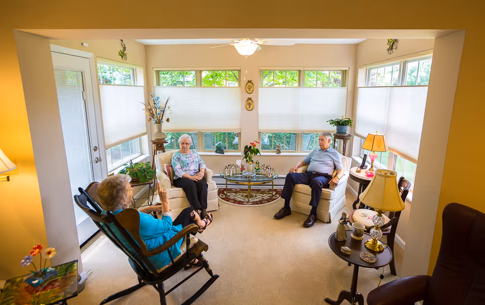 A cozy living room with three elderly people sitting and conversing. The room has large windows with blinds partially drawn, a ceiling fan, and various lamps and plants. There is a glass coffee table with decorative items in the center and comfortable armchairs arranged around it.