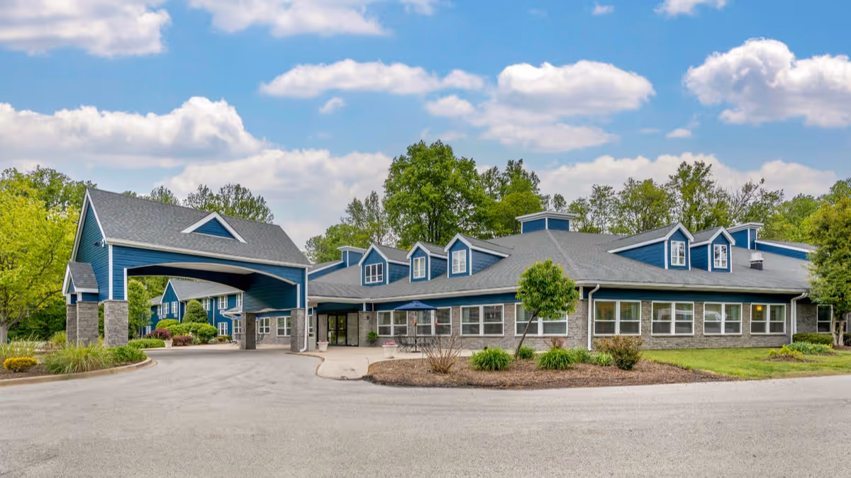 Front exterior of a blue senior living building with a covered porte-cochère entrance, landscaped grounds, and a circular driveway.