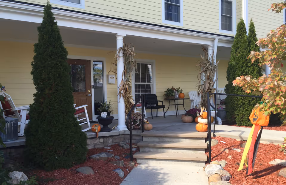 Front porch of a yellow building decorated with pumpkins, cornstalks, and fall-themed decorations. There are white columns, rocking chairs, a small table with chairs, and evergreen shrubs on either side of the steps leading up to the porch.