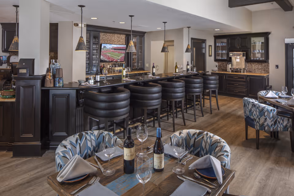 A dining area with a long bar counter featuring black cushioned bar stools and pendant lights hanging above. The bar has bottles of wine and glasses on it. In the foreground, there are tables set with plates, folded napkins, wine glasses, and bottles of wine. The room has wooden flooring and dark cabinetry with glass doors displaying cups and glasses.
