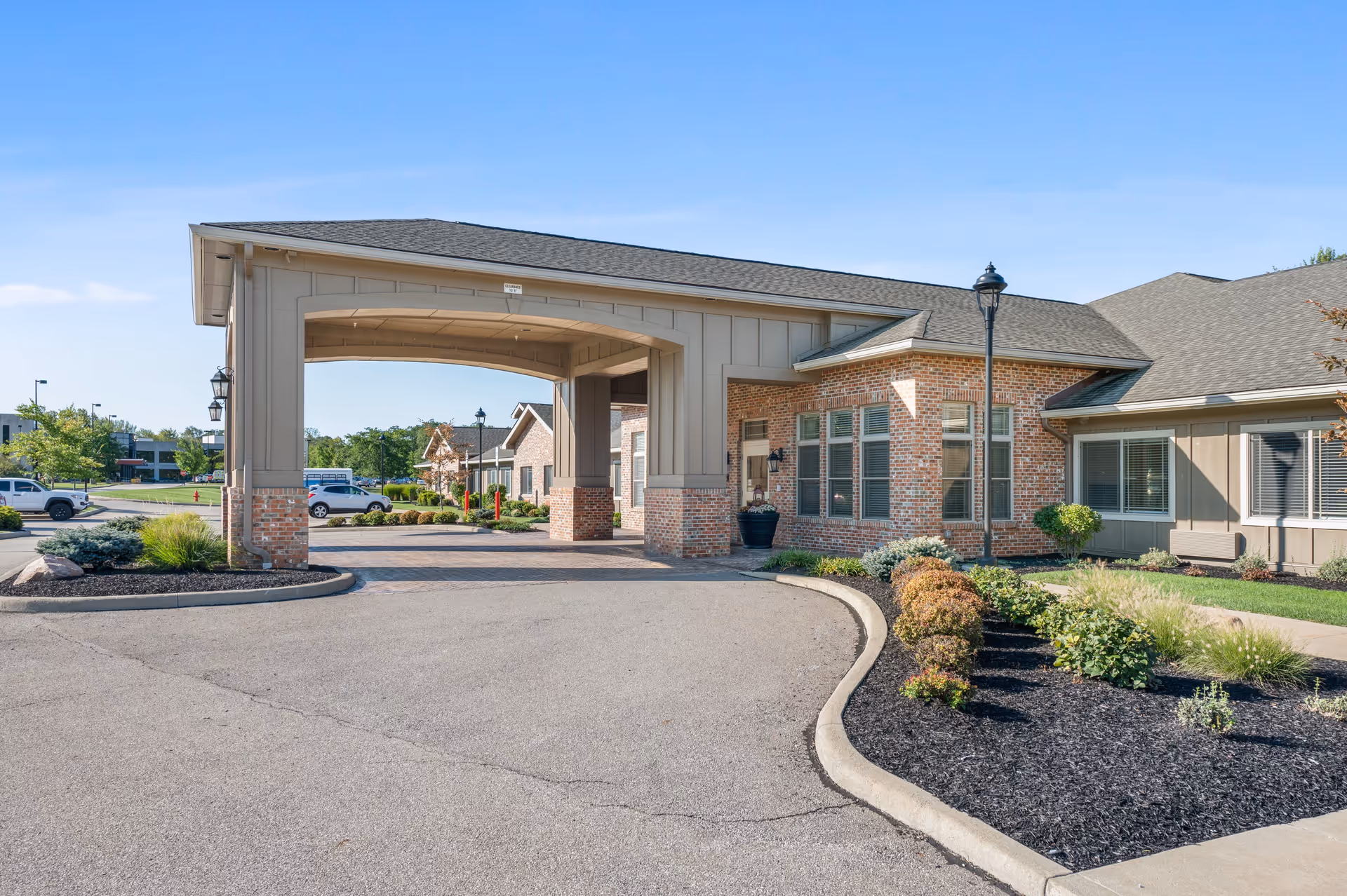 Exterior view of Cardinal Court Assisted Living & Memory Care building entrance with a covered driveway, brick and siding facade, landscaped garden beds, and a clear blue sky.