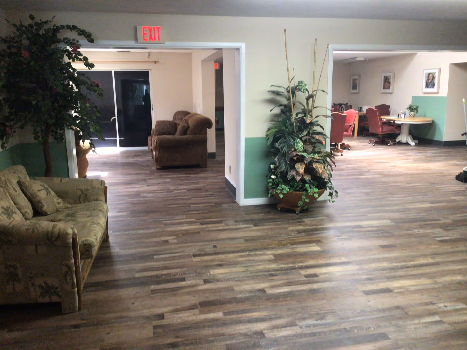 Interior view of a senior living facility showing a spacious area with wood flooring, a green and white painted wall, two couches, a large potted plant, and a dining area with round tables and red chairs in the background. An exit sign is visible above a doorway.