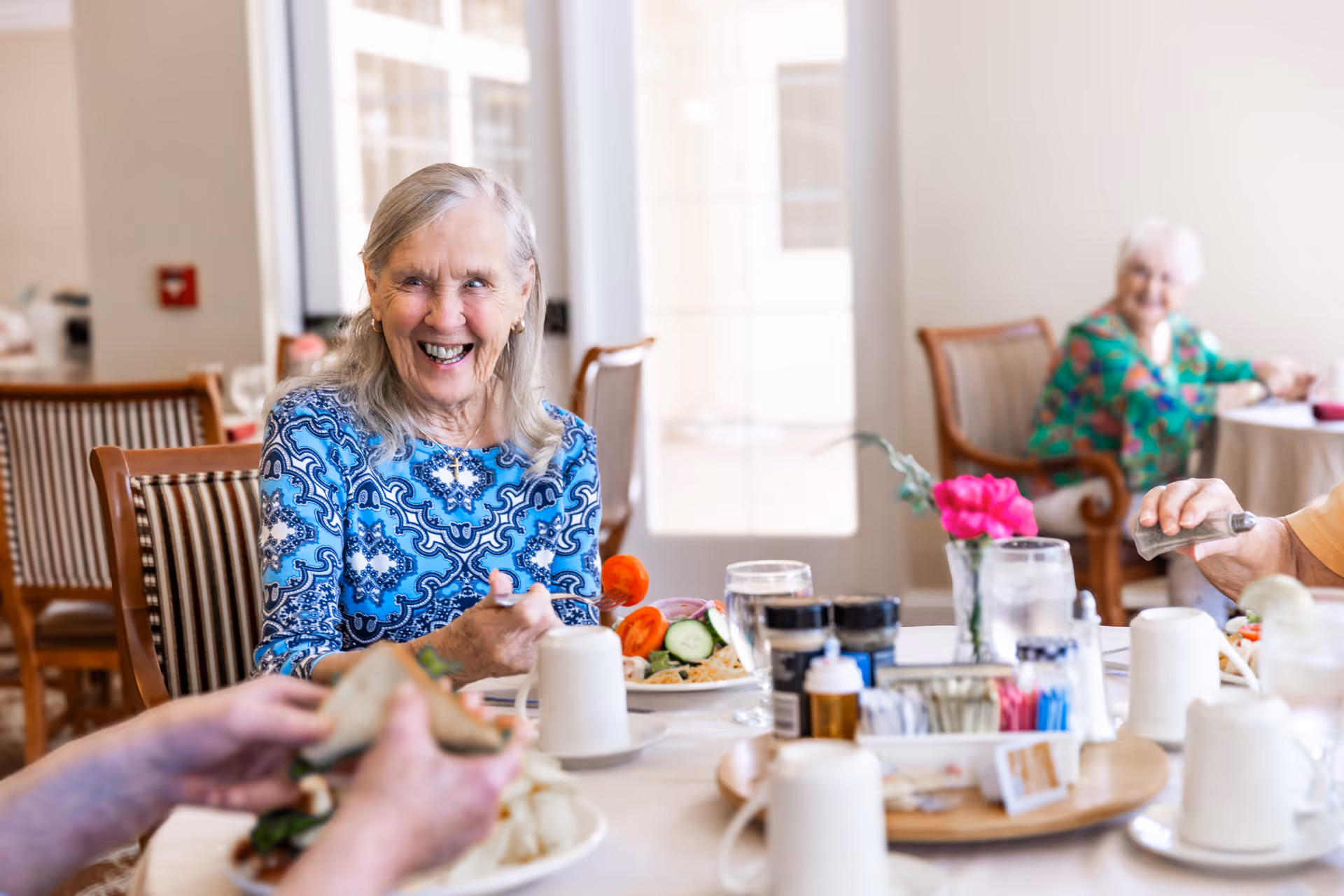 Smiling elderly woman seated at a communal dining table with plates, condiments, and other seniors in the background.