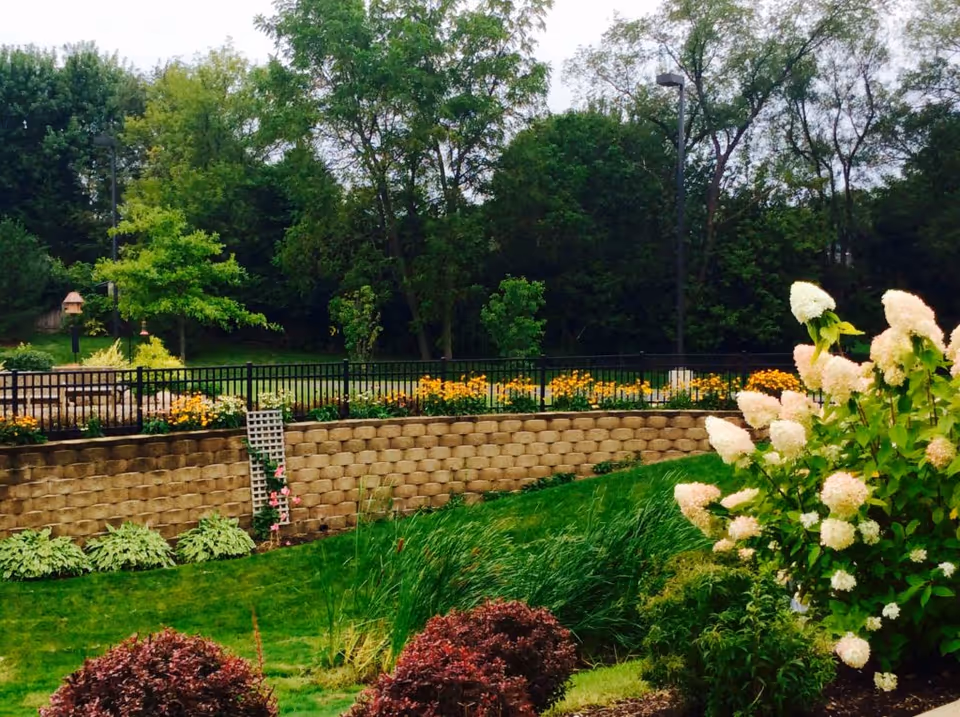 A landscaped outdoor garden area with a stone retaining wall, black metal fence, various flowering plants including white and yellow flowers, green grass, and trees in the background under a cloudy sky.