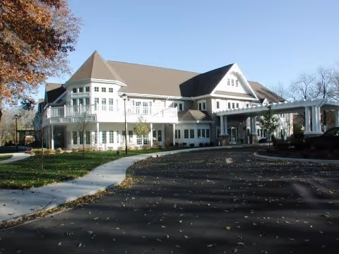 Exterior view of a large, multi-story assisted living facility building with white siding and a brown roof. The building features many windows, a covered entrance with columns, and a curved driveway surrounded by trees with autumn leaves.