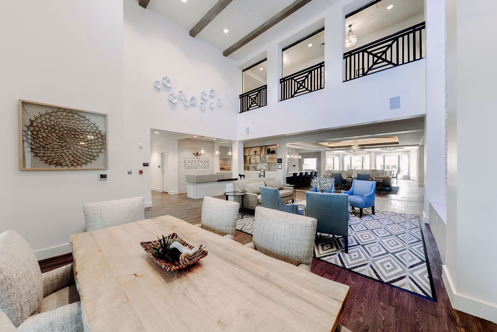 Spacious and modern senior living facility common area with high ceilings and exposed beams. The room features a wooden dining table with upholstered chairs in the foreground, a seating area with blue and beige armchairs on a patterned rug, and a reception desk with the Capstone at Royal Palm Senior Living logo on the wall. Large windows allow natural light to fill the space.