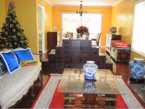 A cozy living room with yellow walls and wooden flooring. The room features a white vintage sofa with blue cushions on the left, a glass-top coffee table with a decorative blue and white vase in the center, and a Christmas tree in the corner. At the back, there is a wooden cabinet with multiple drawers and a dining area with chairs and a table adorned with a flower arrangement. Additional furniture includes a chest with cushions on the right side and another blue and white vase on a stand near the window.