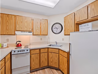 A small kitchen with wooden cabinets, a white stove with a red kettle on it, a white refrigerator, a sink, and a wall clock above the sink. There is a sign on top of the refrigerator that says 'HOME'.