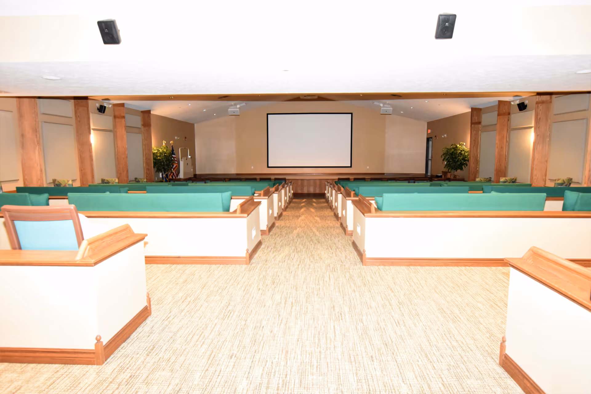 Carpeted meeting room with rows of green-upholstered pews facing a large projection screen at the front.