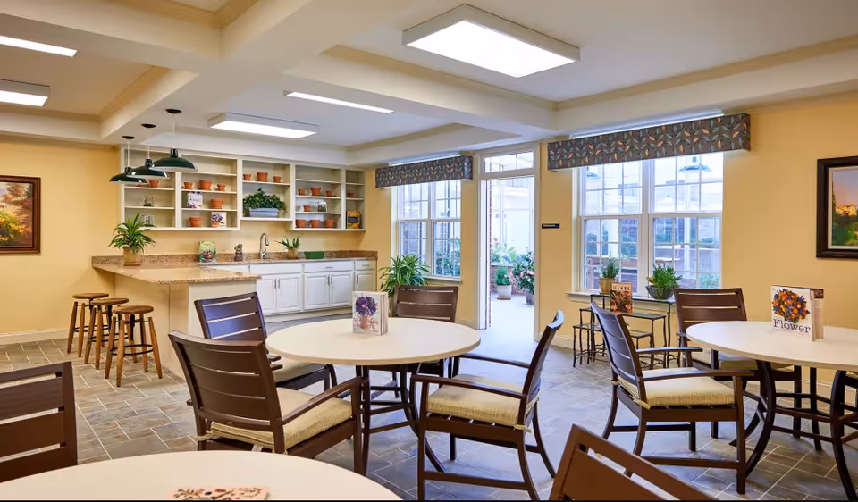 Bright communal dining area with round tables and chairs, a kitchenette counter with stools, and large windows letting in natural light.