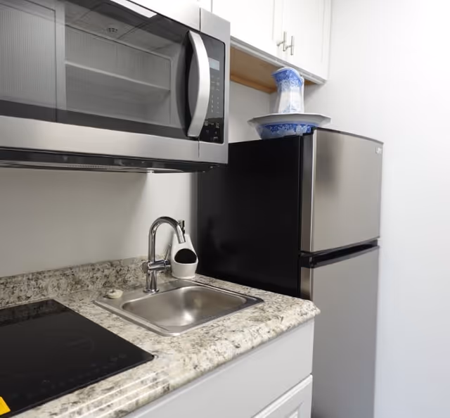 A small kitchen area featuring a stainless steel microwave mounted above a granite countertop with a built-in sink and faucet. Next to the sink is a black electric stovetop. A stainless steel refrigerator is positioned beside the countertop, with a blue and white decorative pitcher and bowl placed on top. White cabinets are visible above the countertop and refrigerator.