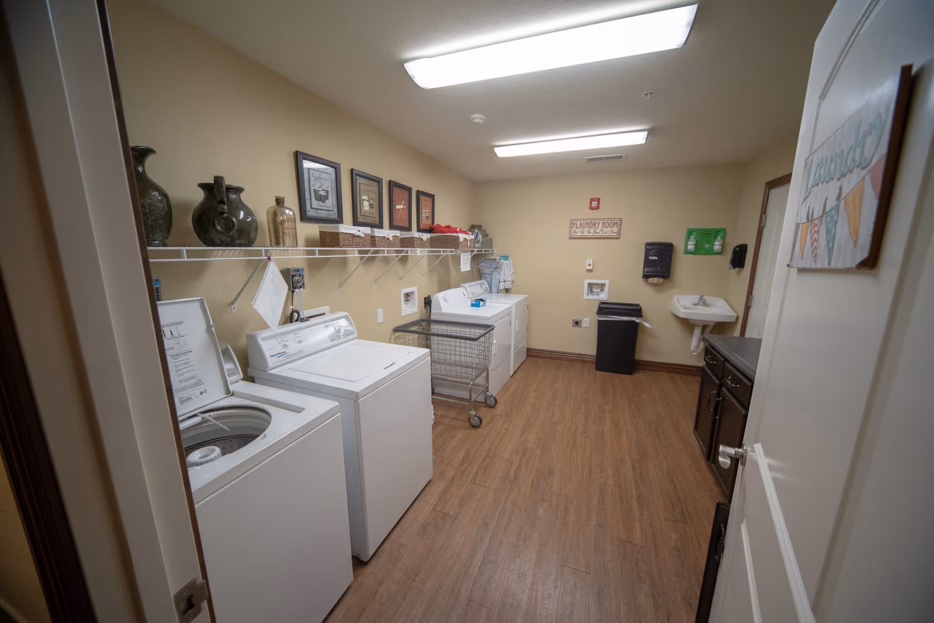 Laundry room with two washing machines and two dryers along the left wall, a laundry cart in the center, a sink and countertop on the right, and decorative items and framed pictures on a shelf above the machines. The room has wood flooring and beige walls with a sign that reads 'Laundry Room'.