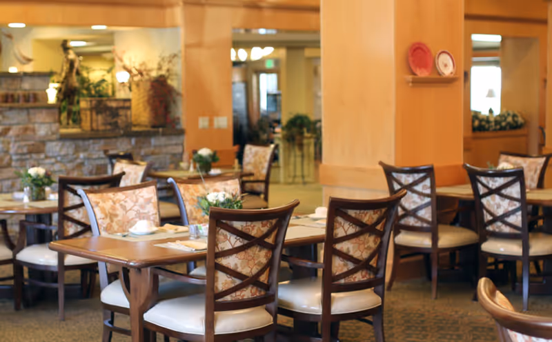 Dining room with wooden tables and upholstered chairs set for meals in a senior living facility.