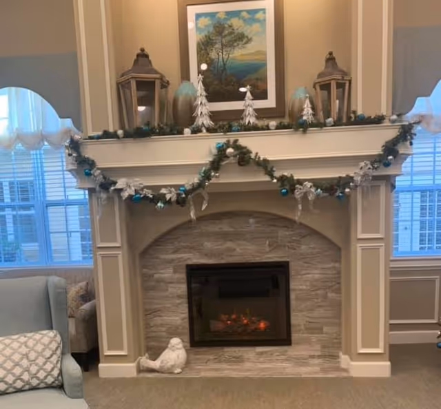 Fireplace mantel decorated with garland, lanterns and ornaments, a framed landscape above, and an electric fire in a living room.