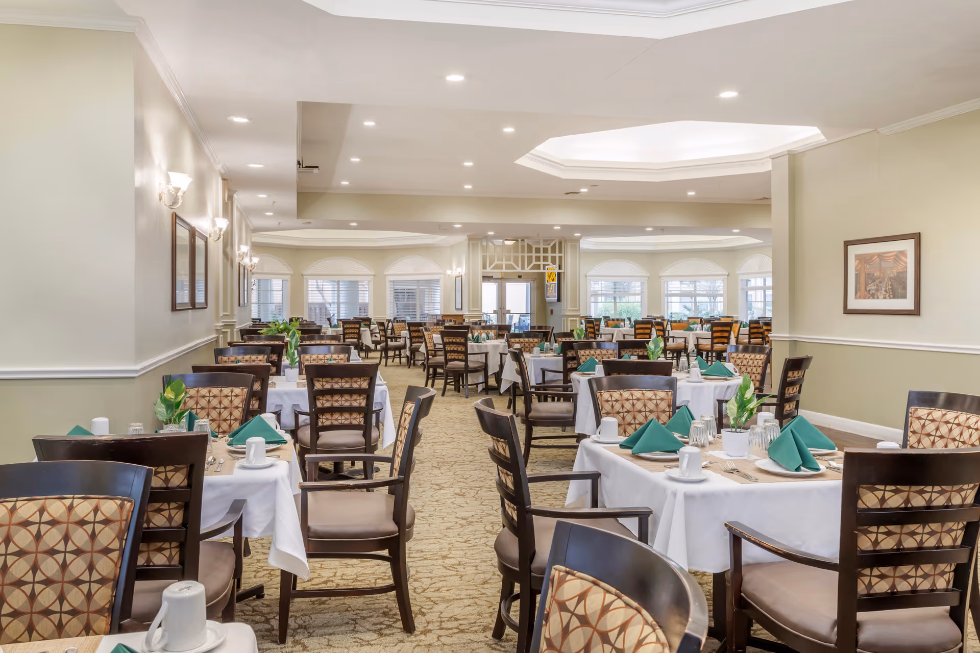A spacious dining room with multiple tables covered in white tablecloths, each set with green folded napkins, cups, glasses, and small potted plants. The room has beige walls, carpeted floors, and large windows letting in natural light. The ceiling features recessed lighting and a decorative tray ceiling.