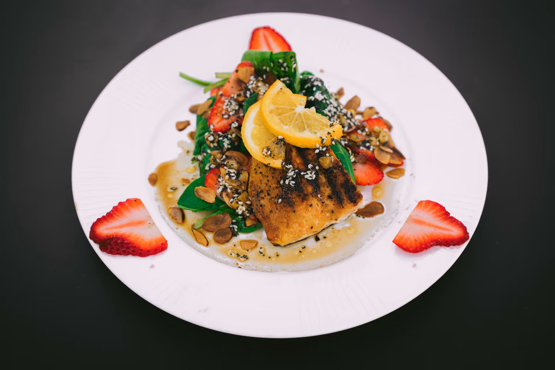 A plated dish featuring a grilled piece of fish topped with lemon slices, served on a bed of spinach and garnished with sliced strawberries, nuts, and sesame seeds on a white plate with a black background.