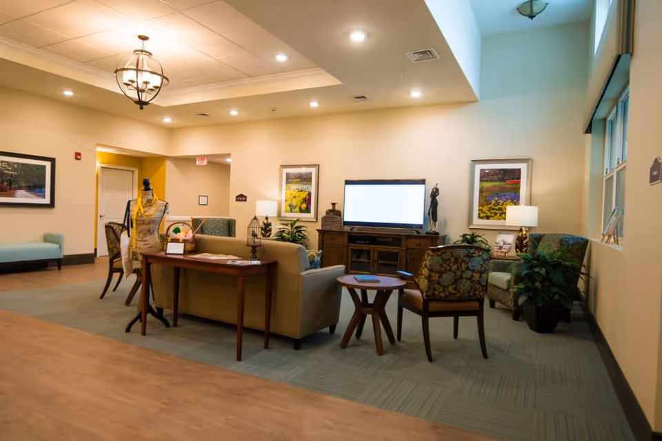 A cozy living room area in a senior living facility with a beige sofa, patterned armchairs, a wooden TV stand with a flat-screen TV, framed floral artwork on the walls, a wooden table with decorative items, and soft lighting from ceiling fixtures and table lamps.
