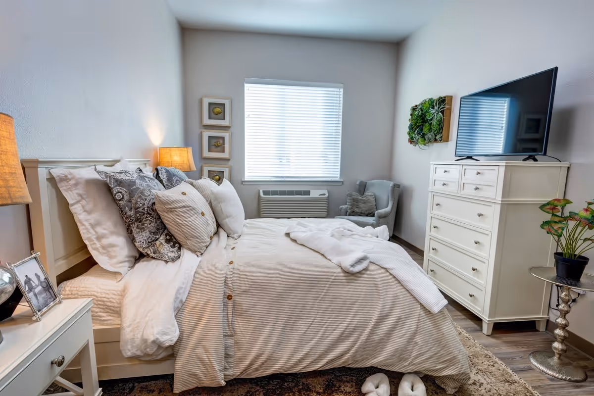 A cozy bedroom with a neatly made bed featuring multiple pillows and a beige striped comforter. There is a bedside table with a lamp and a framed photo, a window with blinds, a gray armchair with a pillow, a white dresser with a flat-screen TV on top, and a small round table with a potted plant. The room has light-colored walls and wooden flooring.