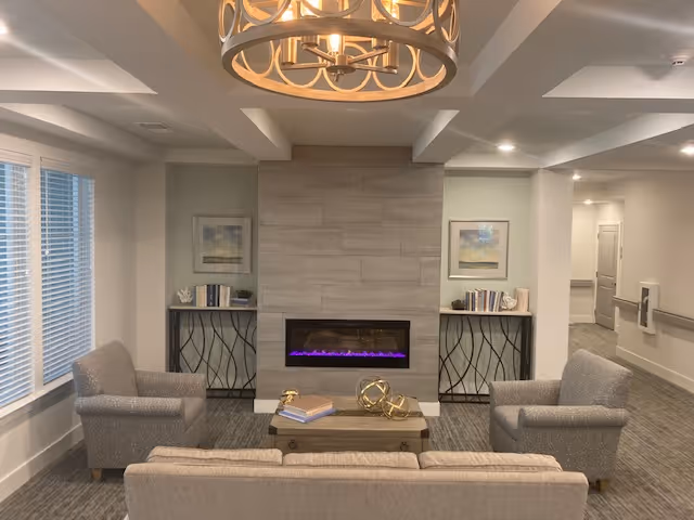 A cozy senior living common area with a modern electric fireplace set in a tiled wall. The room features a beige sofa and two gray armchairs arranged around a wooden coffee table with decorative items. Two side tables with books and decor flank the fireplace, and two framed pictures hang on the wall. Large windows with blinds are on the left, and a stylish chandelier hangs from the ceiling.