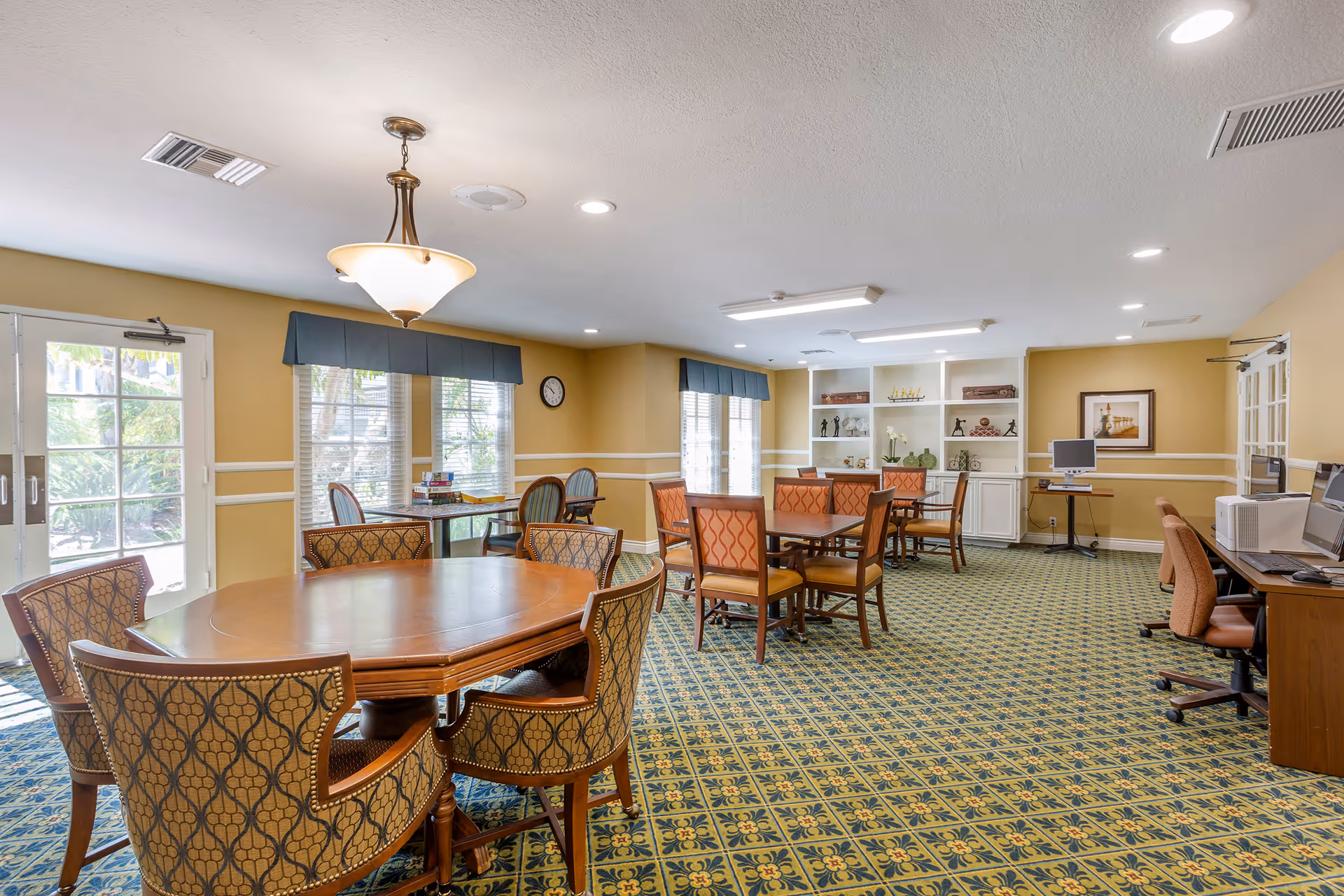 A spacious room with multiple wooden tables and upholstered chairs arranged for group seating. The room has patterned carpet flooring, beige walls with white trim, and large windows with blue valances allowing natural light. There are built-in white shelves with decorative items, a clock on the wall, and a computer workstation with chairs along one side. A glass door leads outside to a garden area.