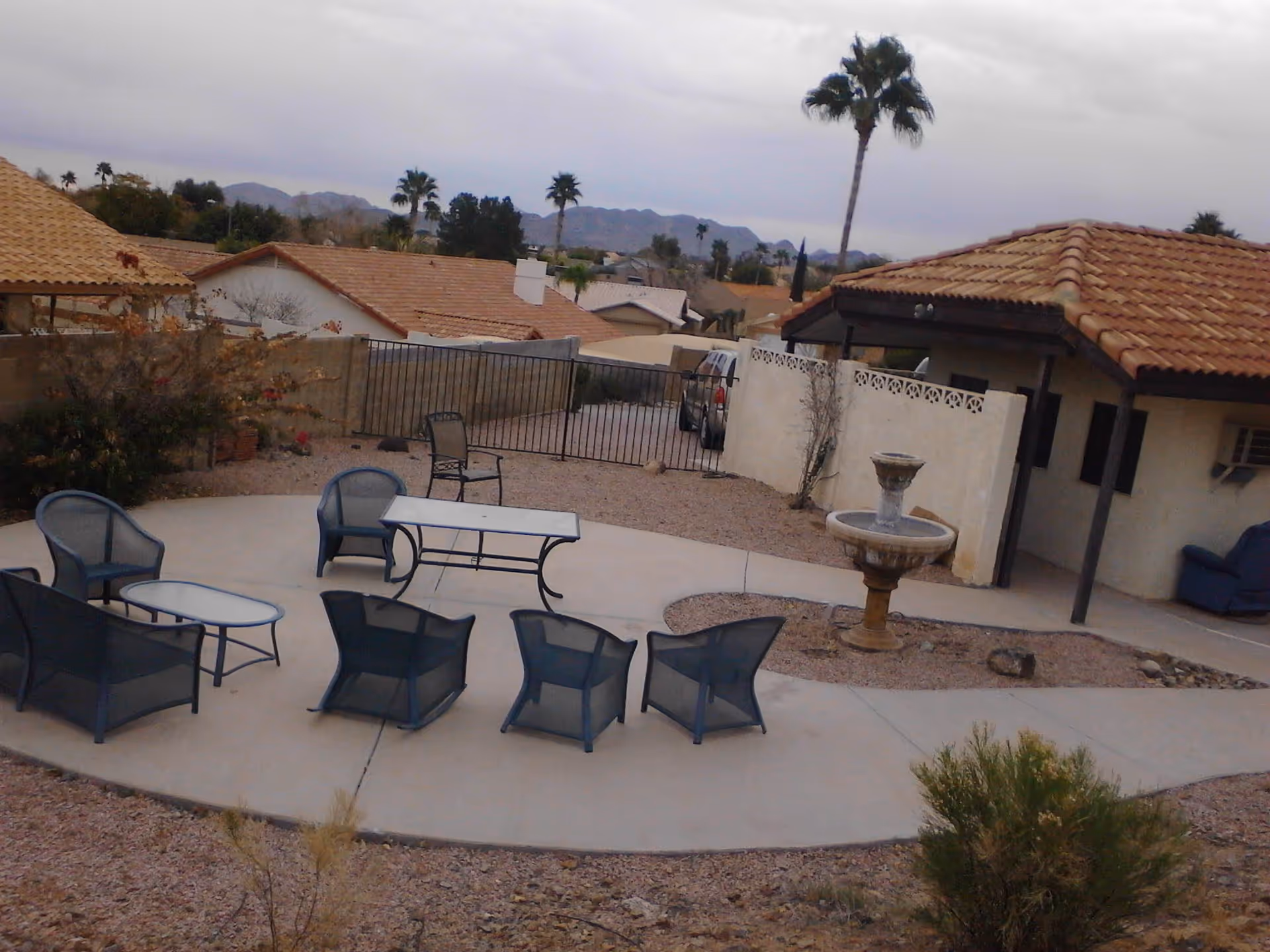 Outdoor patio area with several black mesh chairs and tables arranged on a concrete surface. There is a two-tiered stone fountain on a gravel section near the patio. Surrounding the patio are desert landscaping with gravel and sparse bushes. In the background, there are houses with tiled roofs, palm trees, and mountains under a cloudy sky.