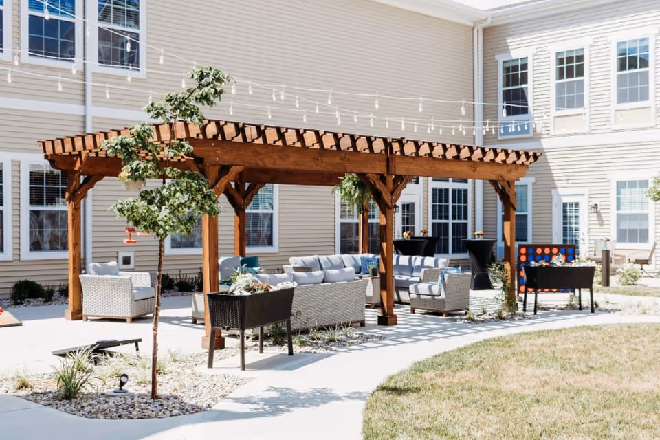 Outdoor courtyard with a wooden pergola, string lights, and cushioned patio seating in front of a multi-story facility building.