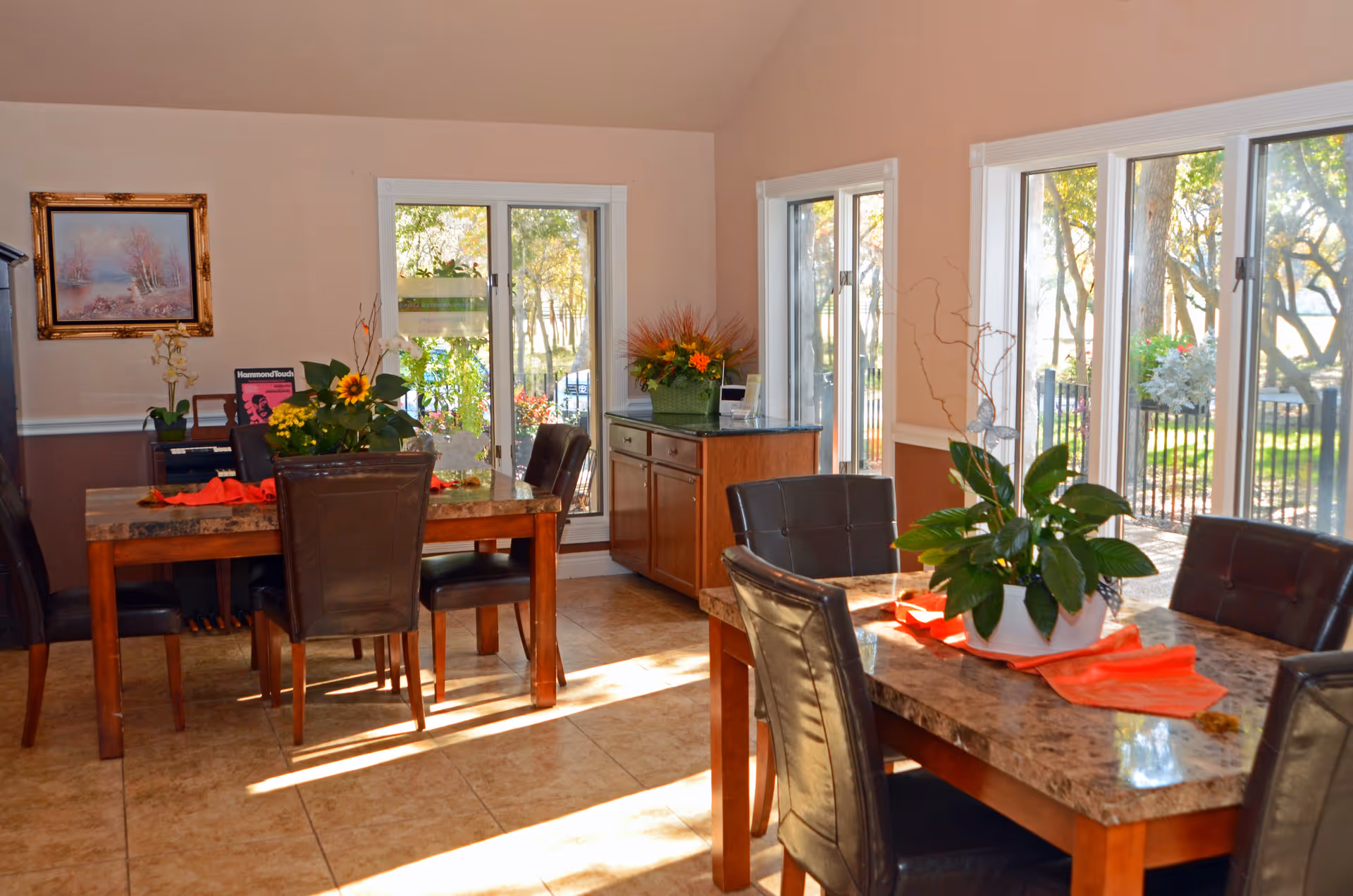 A bright dining room with two marble-top tables surrounded by dark leather chairs. Each table has a centerpiece with green plants and orange napkins. Large windows let in natural light and offer a view of trees and greenery outside. A wooden cabinet with a flower arrangement is against the wall, and a framed painting hangs on the left wall.