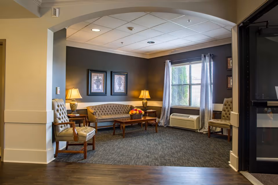 A cozy seating area in a senior living facility with a tufted sofa, two armchairs, a wooden coffee table with a flower arrangement, two table lamps on side tables, framed artwork on the dark accent wall, a window with curtains, and a carpeted floor under the furniture.