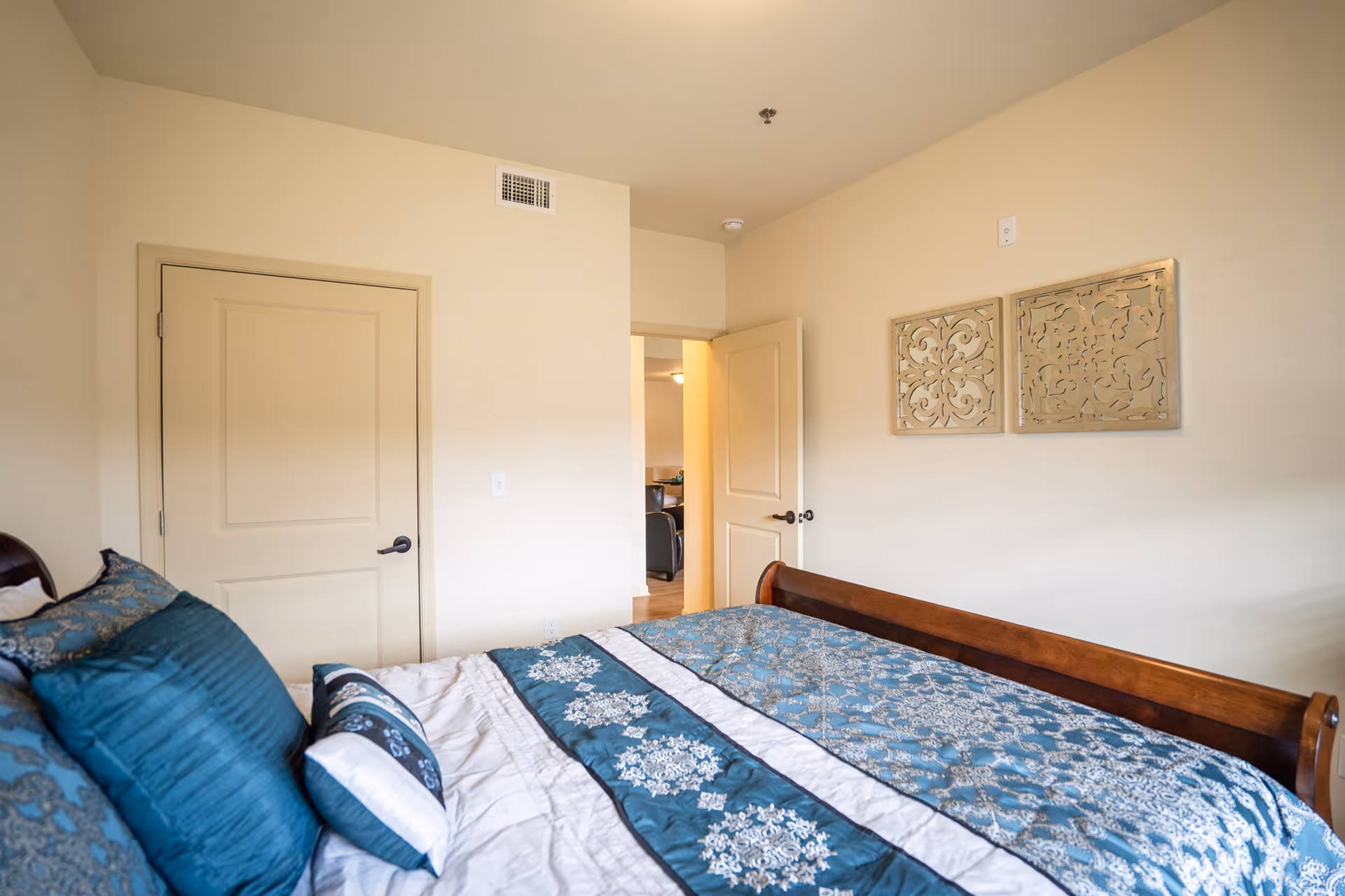 Well-lit bedroom with a wooden sleigh bed dressed in blue patterned bedding, beige walls, and decorative wall panels above the bed.