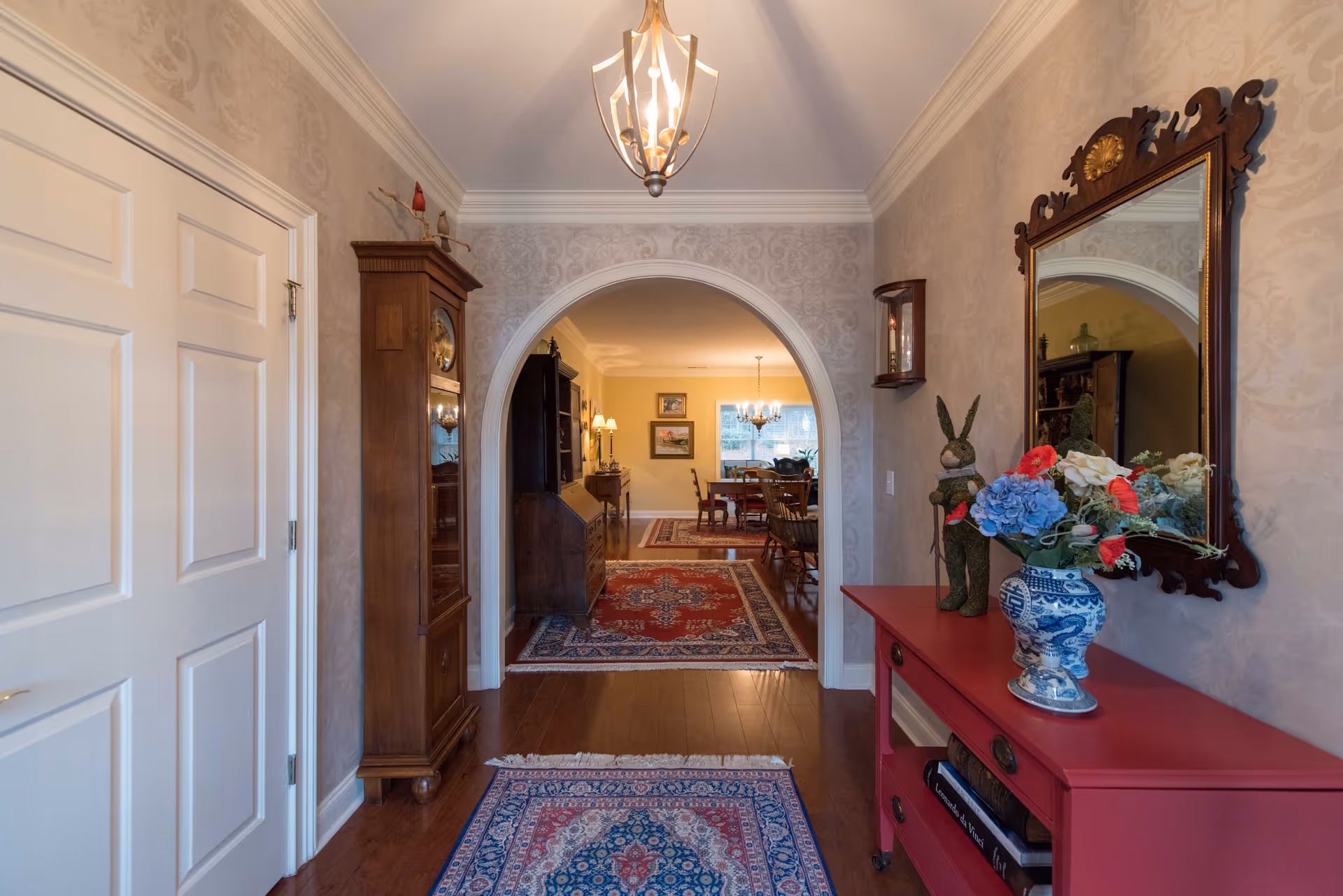 View of an elegant hallway with patterned wallpaper, a red console table holding a blue and white vase with flowers, a decorative rabbit figure, and a large ornate mirror on the right wall. The hallway has wooden floors with a blue and red patterned rug and leads through an arched doorway into a dining room with wooden furniture and a chandelier.