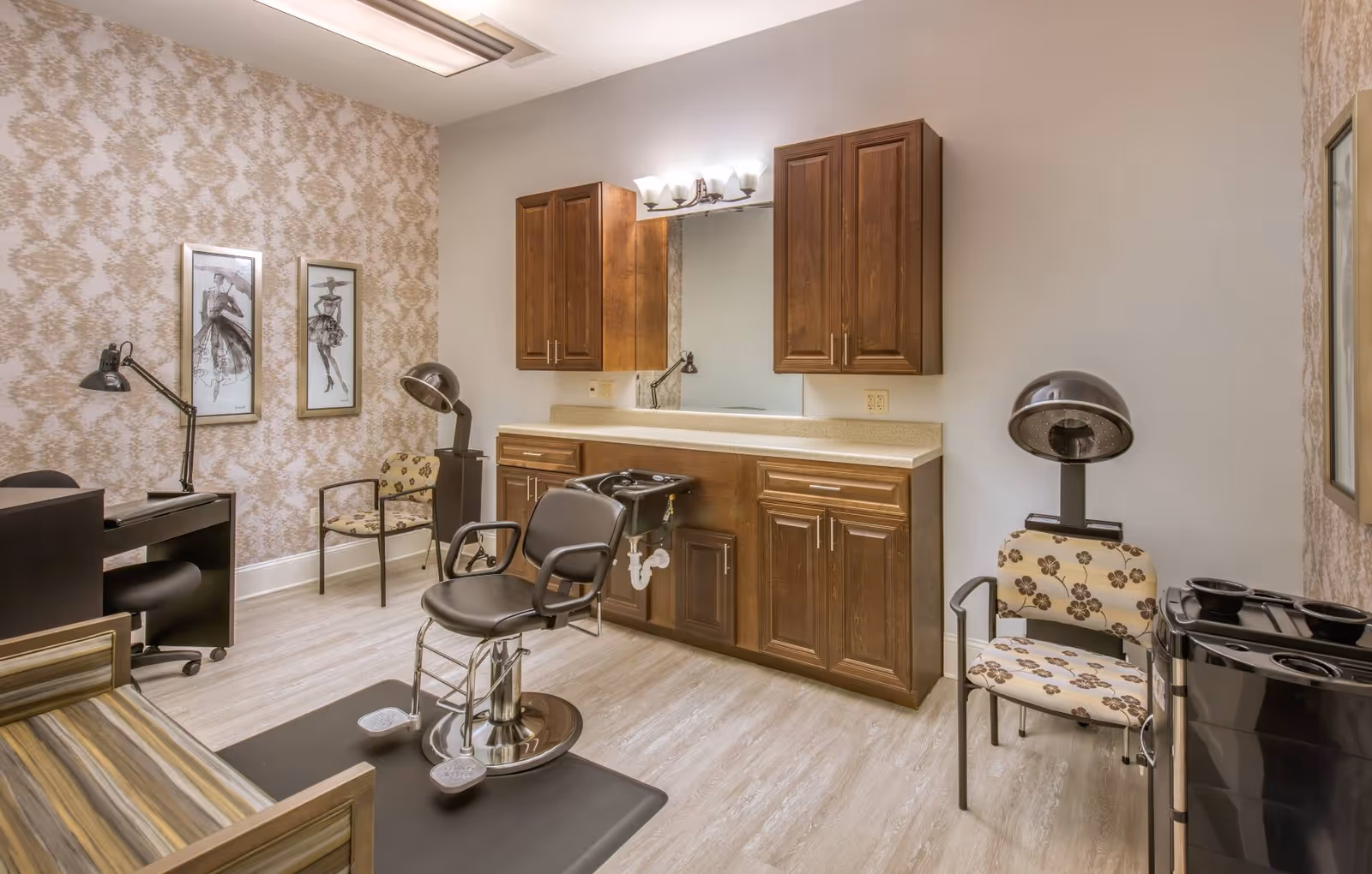 Interior view of a salon room with a styling chair in front of a counter with a sink and wooden cabinets. The room has patterned wallpaper on one wall, two framed fashion sketches, a desk with a lamp, two chairs with floral upholstery, and a hair dryer chair.