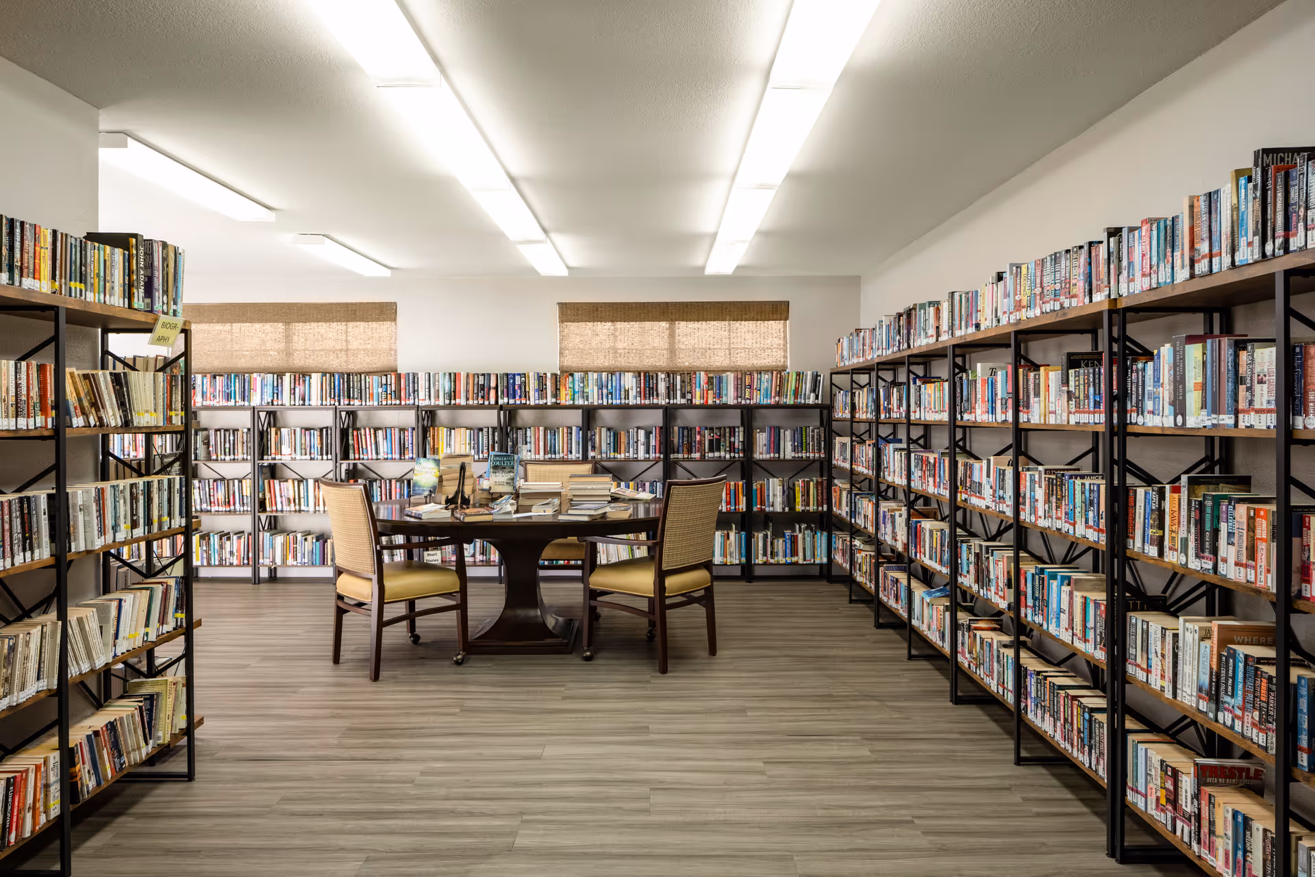 A well-lit library room with multiple bookshelves filled with books along the walls and a round wooden table with four chairs in the center. The floor has a light wood finish, and there are two windows with woven shades on the back wall.