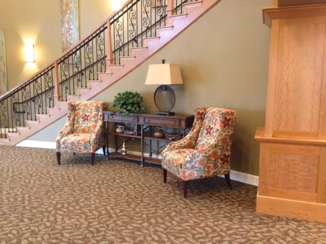 A cozy interior seating area with two floral-patterned armchairs placed on a patterned carpet. Between the chairs is a wooden console table with decorative items and a lamp. Behind the seating area is a staircase with an ornate black metal railing and beige walls with wall-mounted lights.