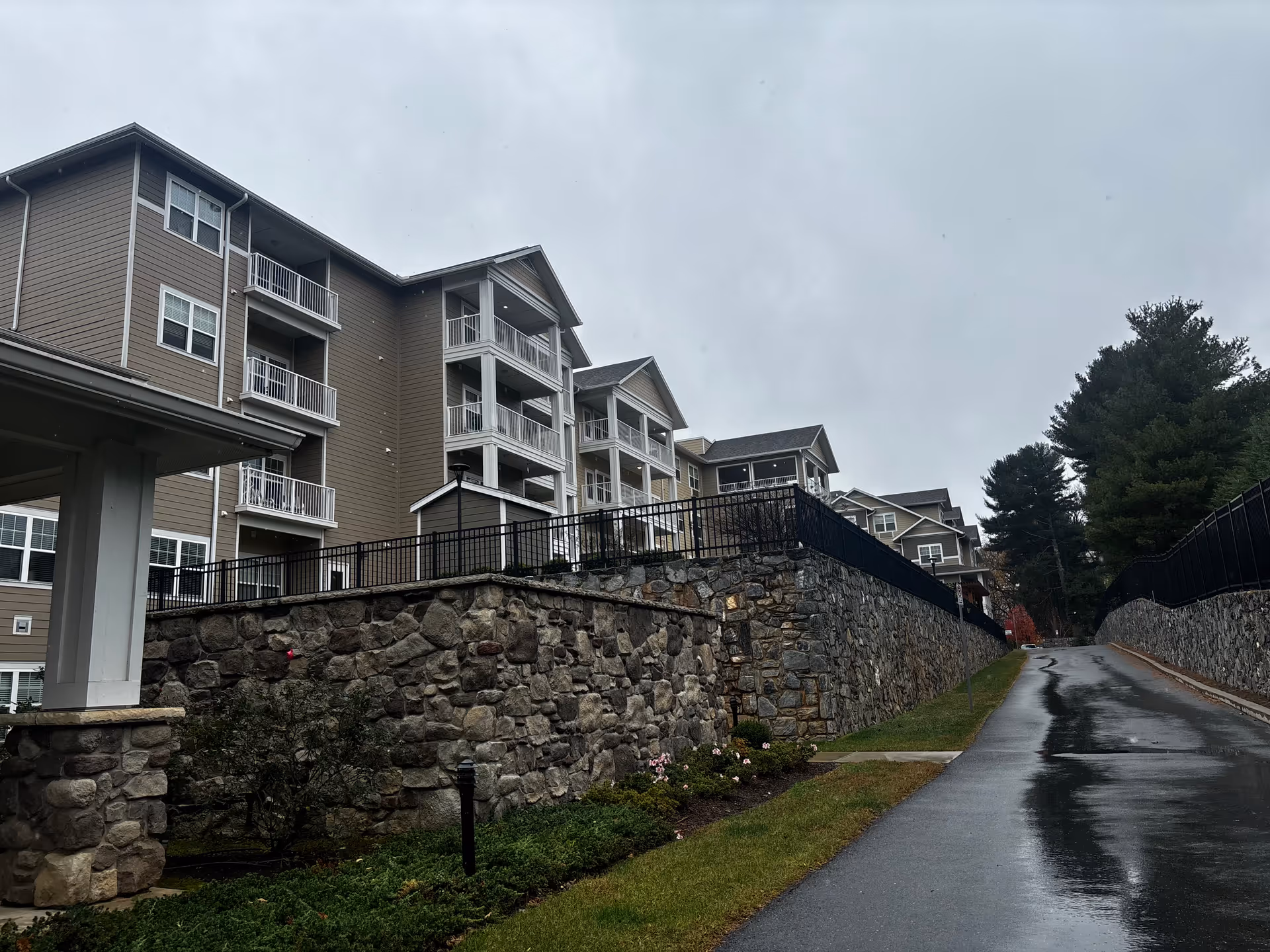 Multi-story senior living building with balconies above a stone retaining wall and a wet driveway leading uphill.